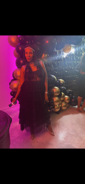FashionTechNora wearing a black-and-white tiered tulle dress with a sheer structured corset bodice, pearl headband, and pointed snakeskin heels at her son’s grandmother’s 80th birthday celebration. Posed in front of black and gold balloon décor, the monochrome ensemble layers contrasting textures, sheer details, and statement accessories for an elegant editorial-inspired evening look.