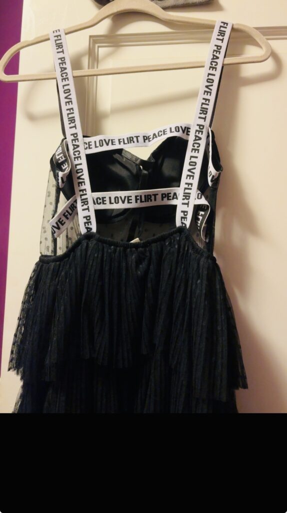 Detail shot of a black dress with “Flirt Peace Love” statement straps hanging on a wooden hanger, featured in FashionTechNora’s “A Love Letter to Black & White” editorial celebrating timeless monochrome style.